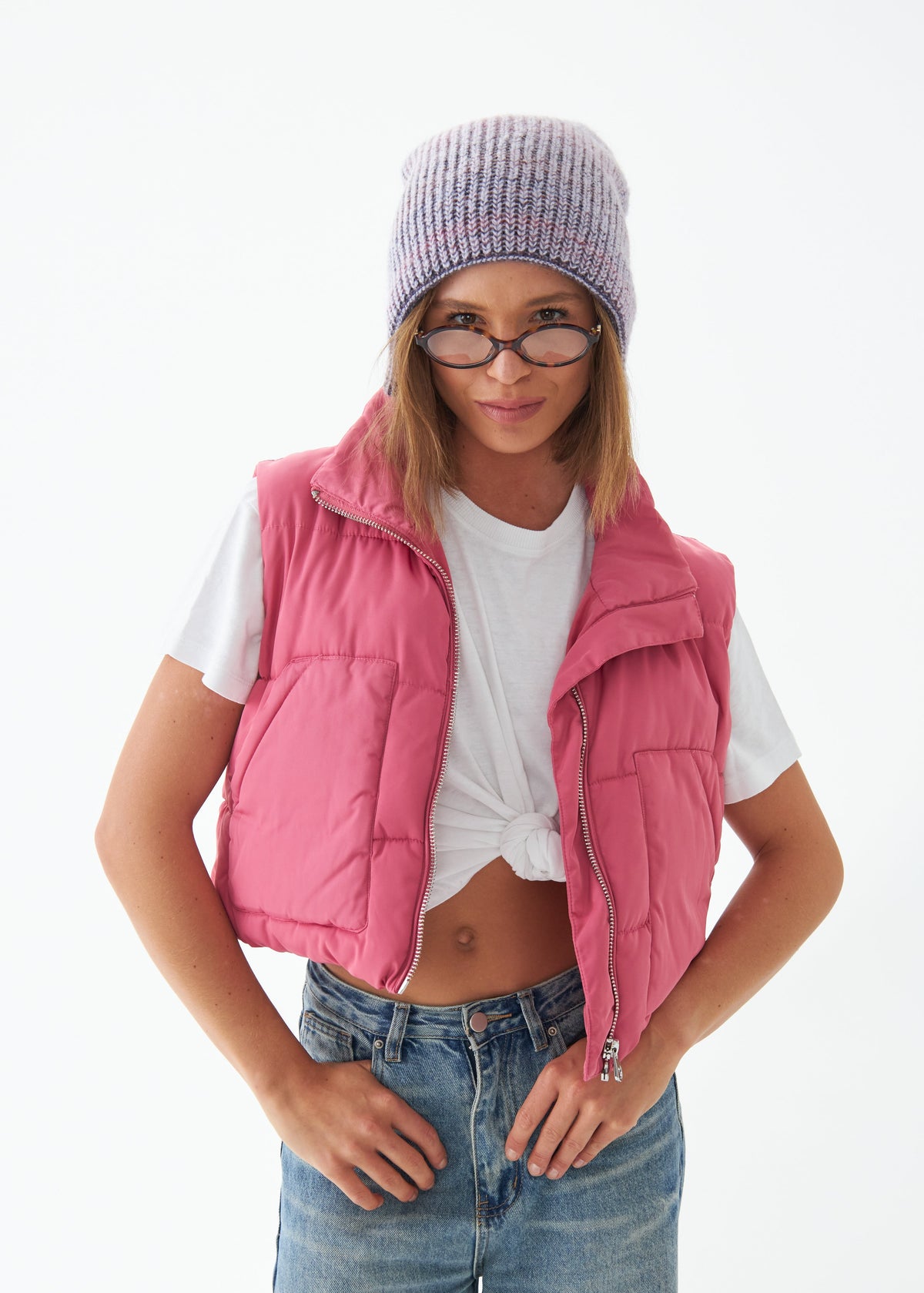 Person wearing a pink puffer vest, white t-shirt, blue jeans, and a gray knit beanie on a white background