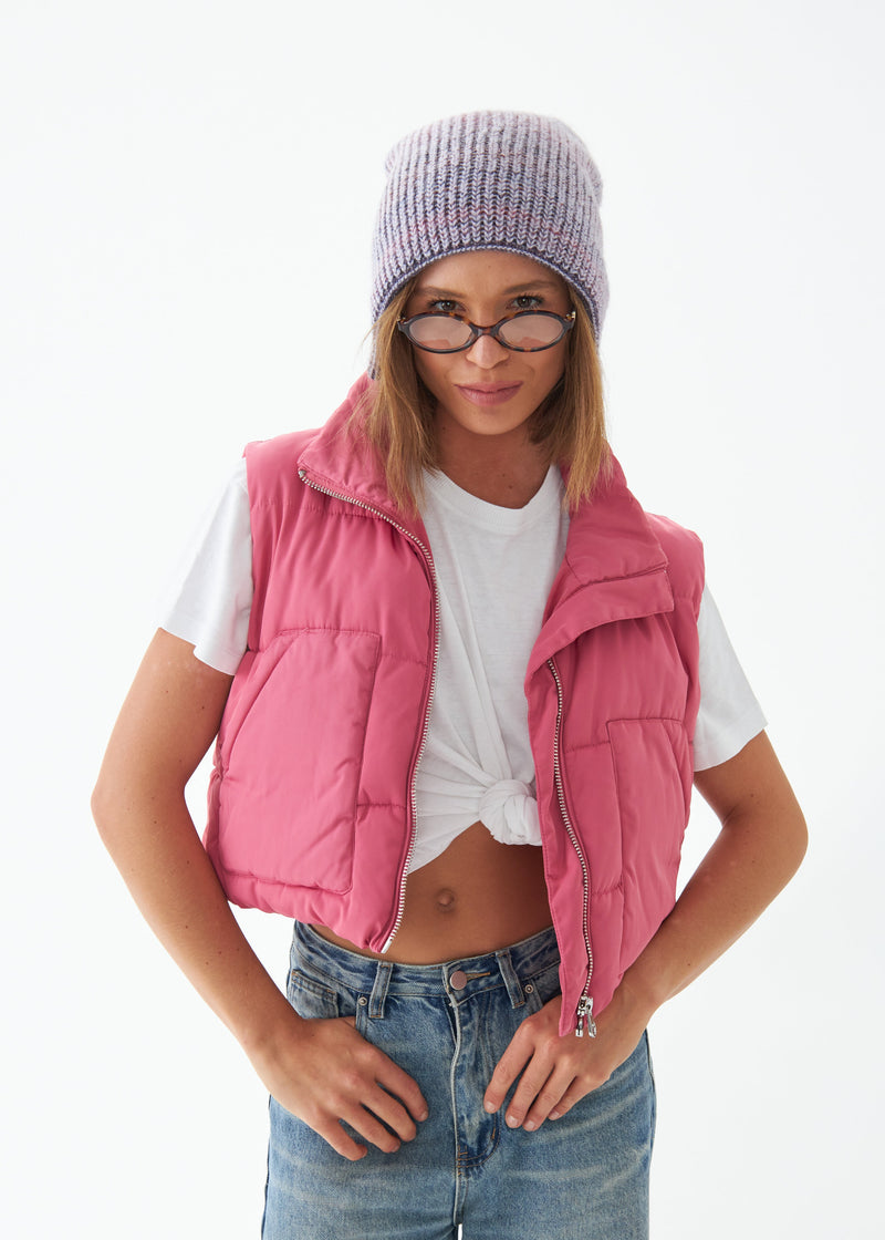 Person wearing a pink puffer vest, white t-shirt, blue jeans, and a gray knit beanie on a white background