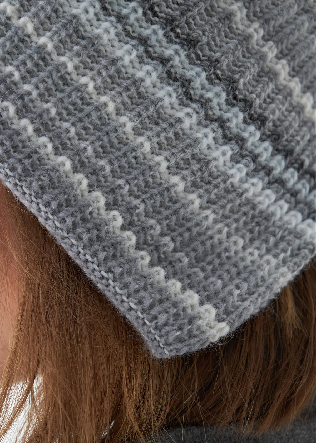 Close-up of a gray knitted beanie with white stripes worn by a person with brown hair.
