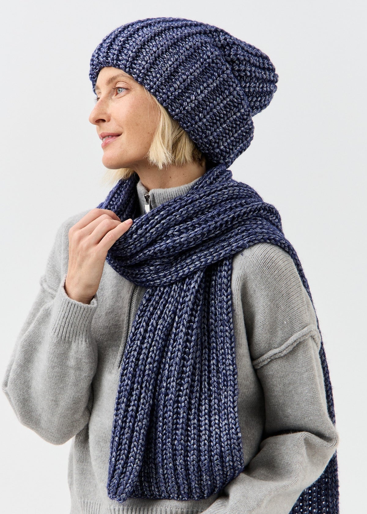 BRIKOLY Lustre Denim Melange oversized knit beanie worn by a model holding a matching scarf near the collar.