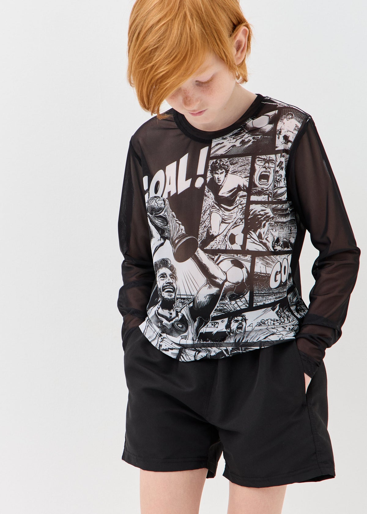 A child wearing a black long-sleeve athletic top with a black-and-white comic-style graphic print and black shorts, photographed against a light gray studio background.