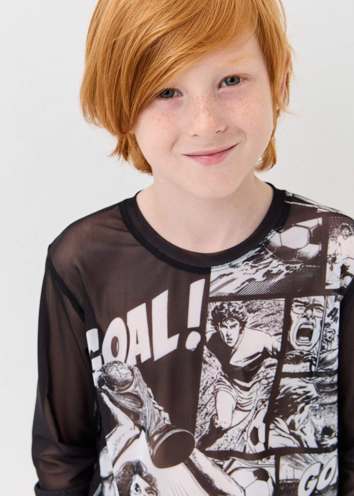 A young boy  wearing a sheer black long-sleeve shirt with a black and white soccer graphic print and the word 'GOAL!' on the front, against a plain light gray background.