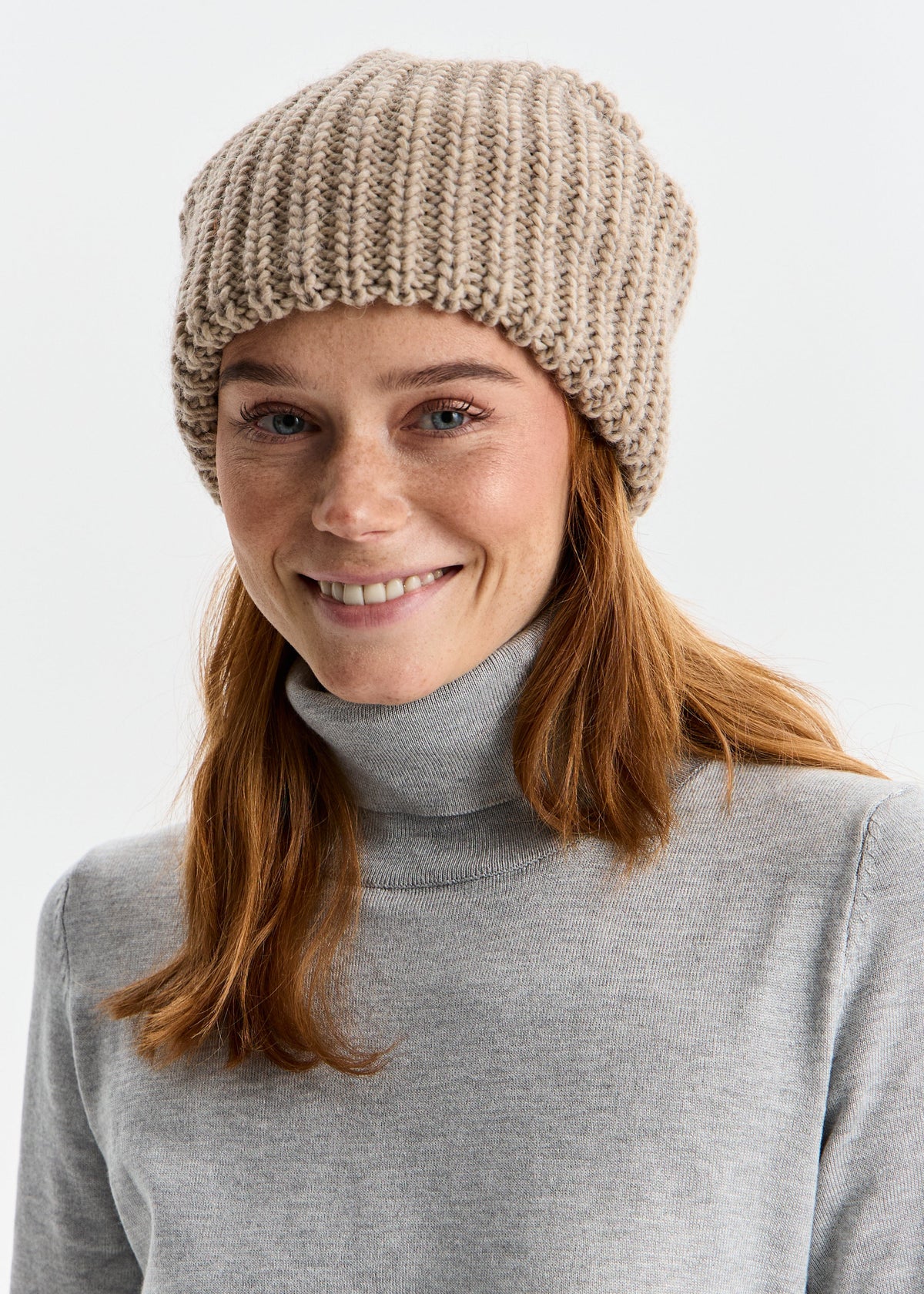 Woman wearing a beige knitted beanie and gray turtleneck against a white background