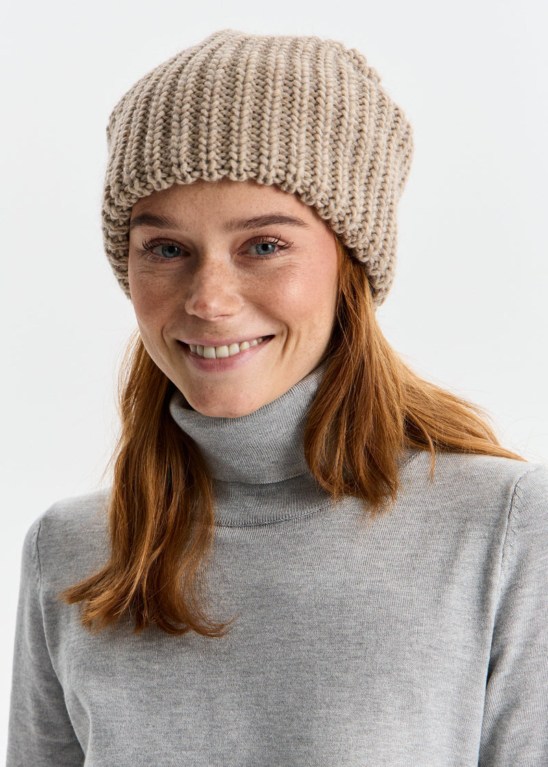 Woman wearing a beige knitted beanie and gray turtleneck against a white background